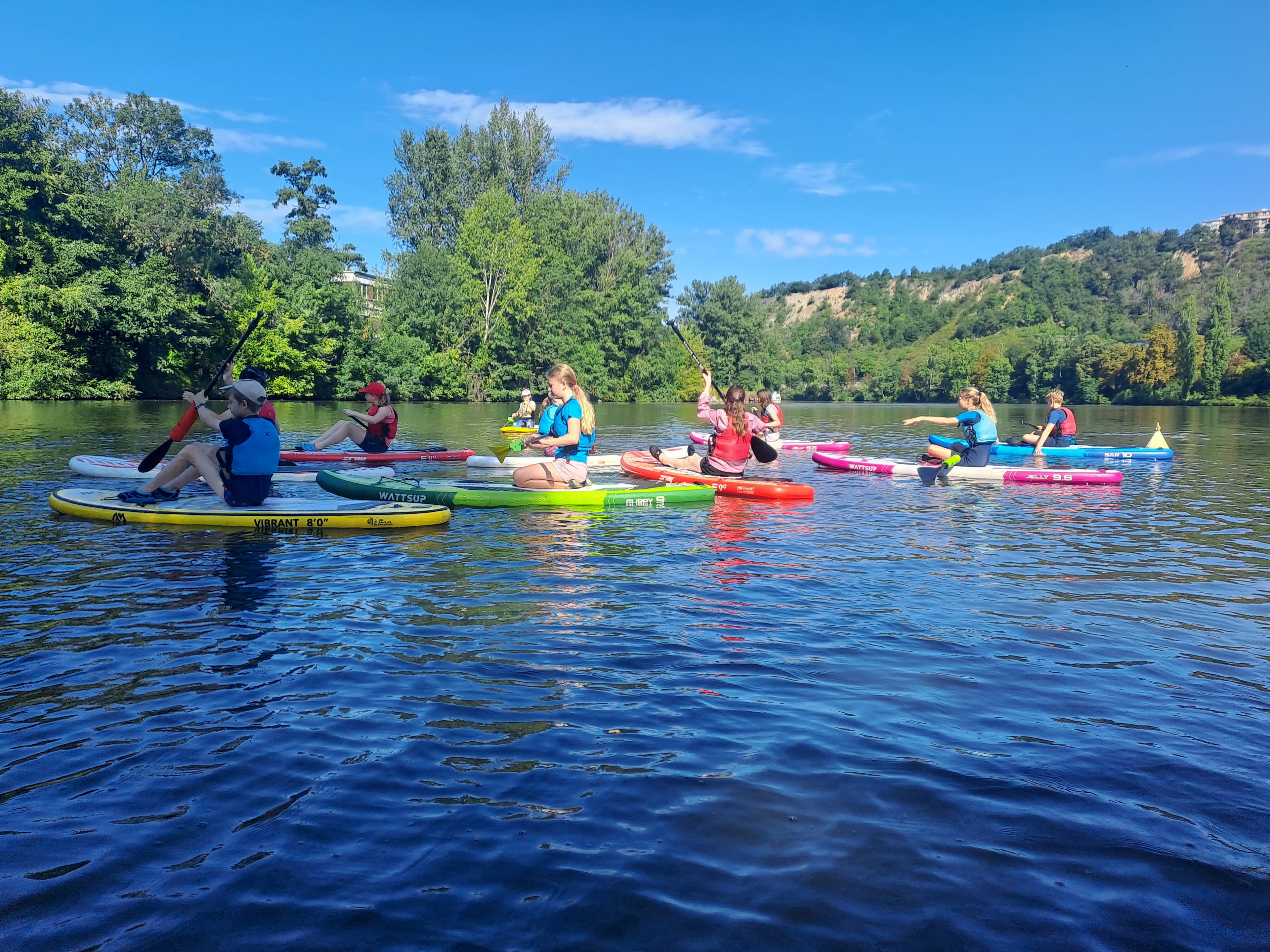 Účastníci vodního programu v záchranných vestách pádlují ve skupině na barevných paddleboardech po Vltavě, v pozadí zalesněné břehy řeky.